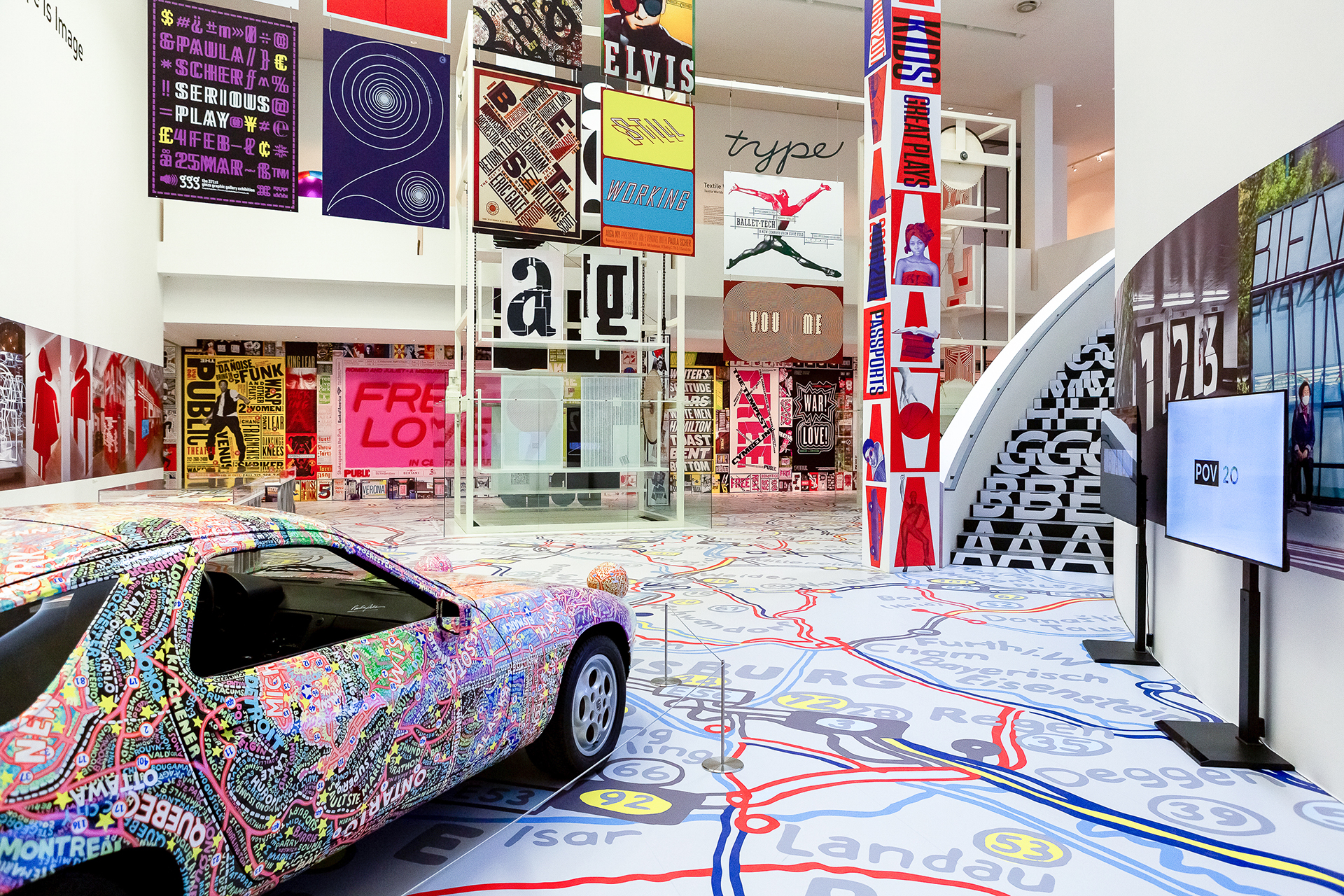 Photo exhibition. In the foreground a car (Porsche), colourfully painted. In the background posters and layouts by Paula Scher. Screens on the right. The floor is a colourful map.