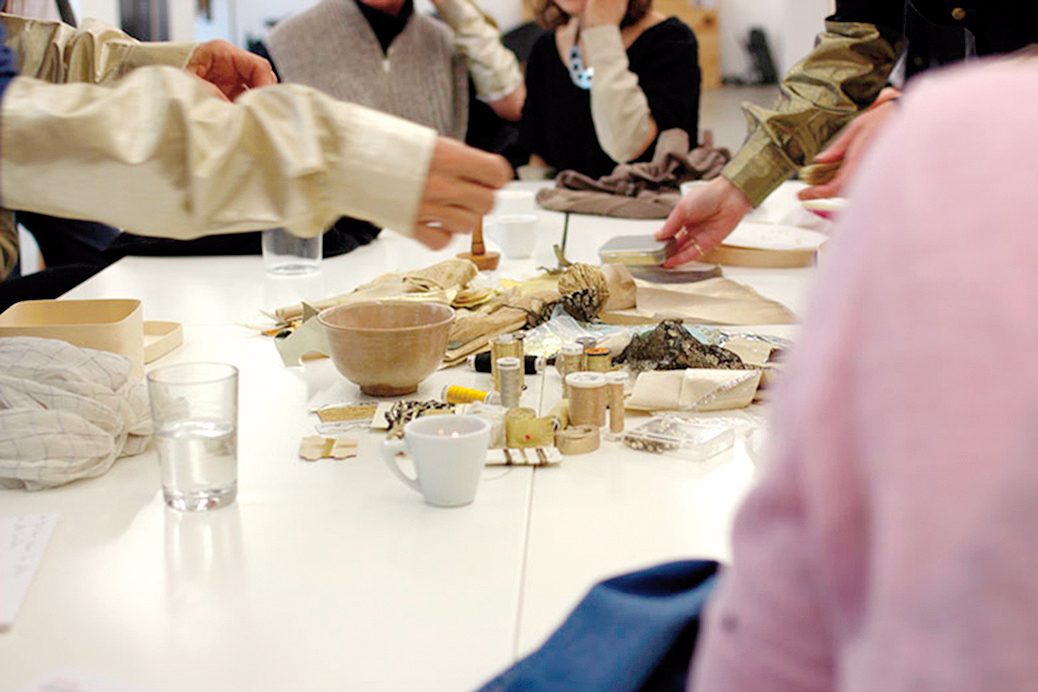 Materials such as gold threads from the "Golden Joinery" workshop lie in the centre of a table. Hands reach for the materials.