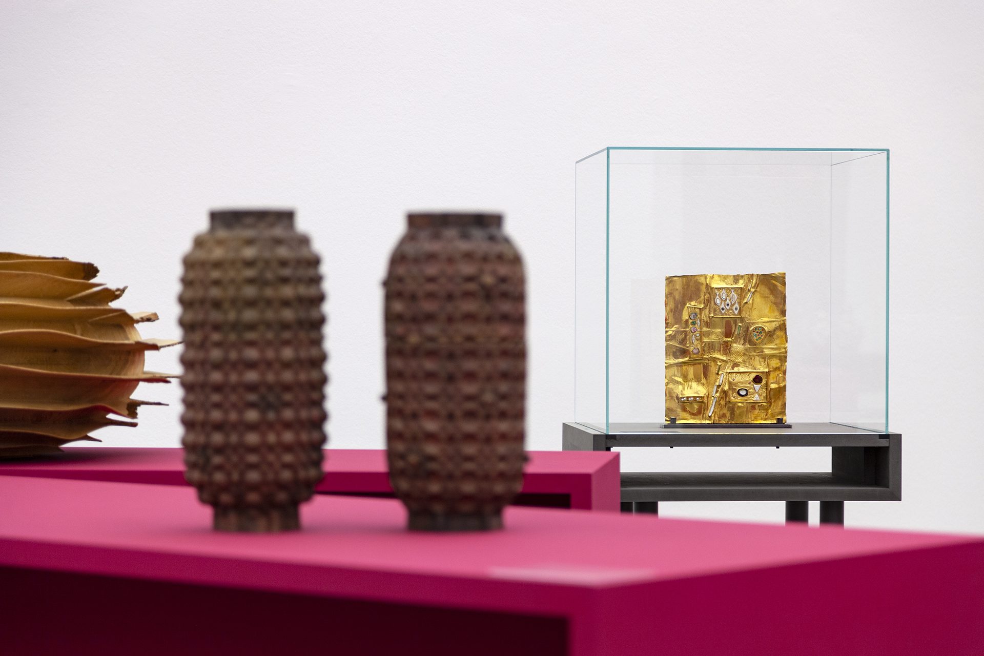 View of the exhibition "Danner Prize 2020. 100 years of the Danner Foundation", red and purple columns in the room, the objects are displayed on pink pedestals/showcases. In the background, design for a tabernacle door by Hermann Jünger, gold-coloured.