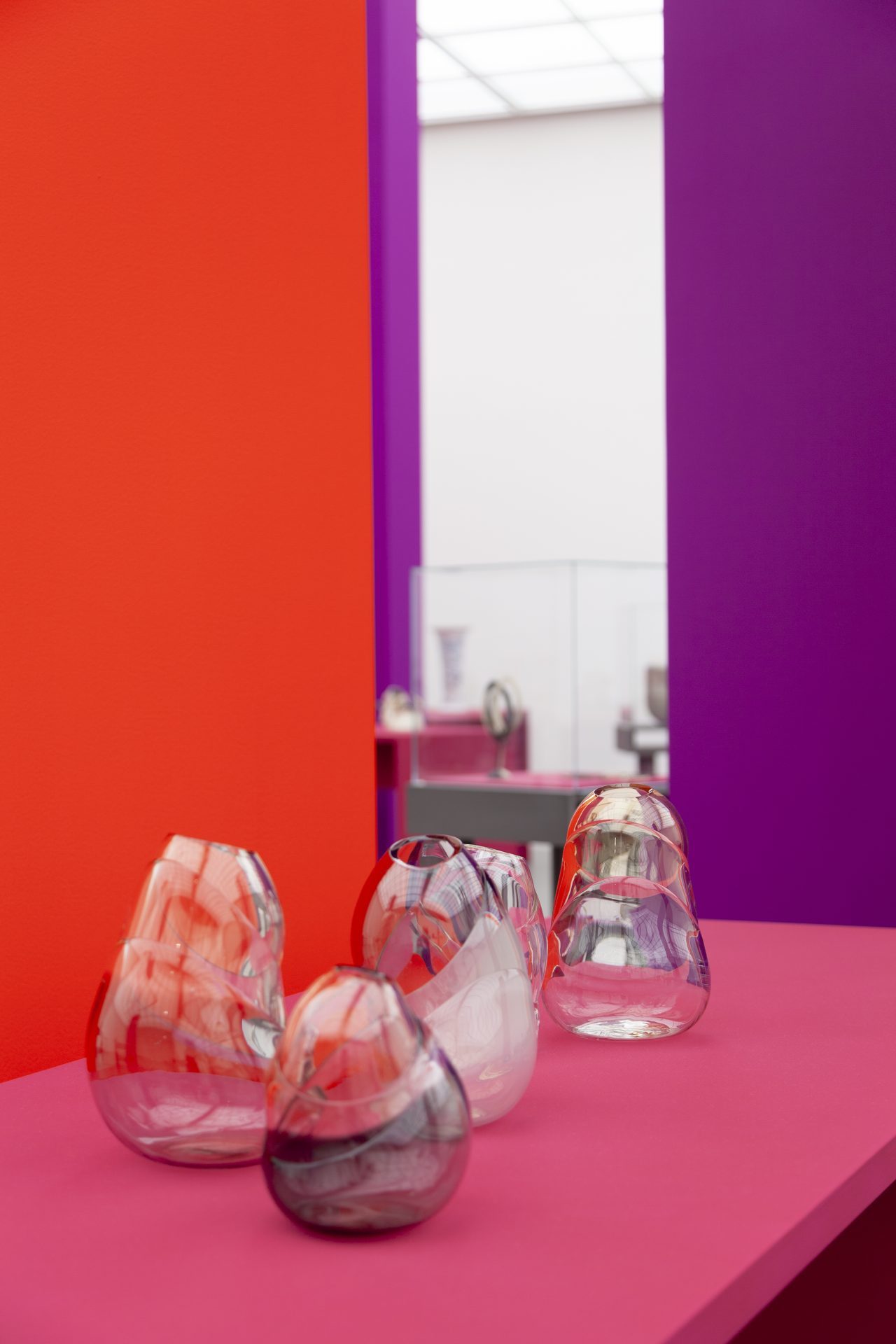 View of the exhibition "Danner Prize 2020. 100 years of the Danner Foundation", red and purple columns in the room, the objects are displayed on pink pedestals/showcases. On display are glass objects by Veronika Beckh. Organically shaped.