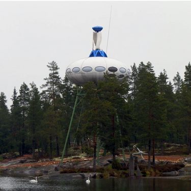 Futuro auf hohen Pfeilern am Rande eines Waldes und Gewässers in Finnland. An der Spitze ragt eine Art blaue, übergroße Antenne aus dem Futuro.