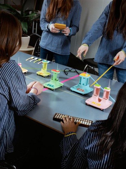 The "Digitarians" in blue and white striped shirts operate the "Digicars", each with 1 or 2 "towers" in the colours light blue, green, pink and yellow, using a remote control. They are labelled with numbers and letters, e.g. H4N.