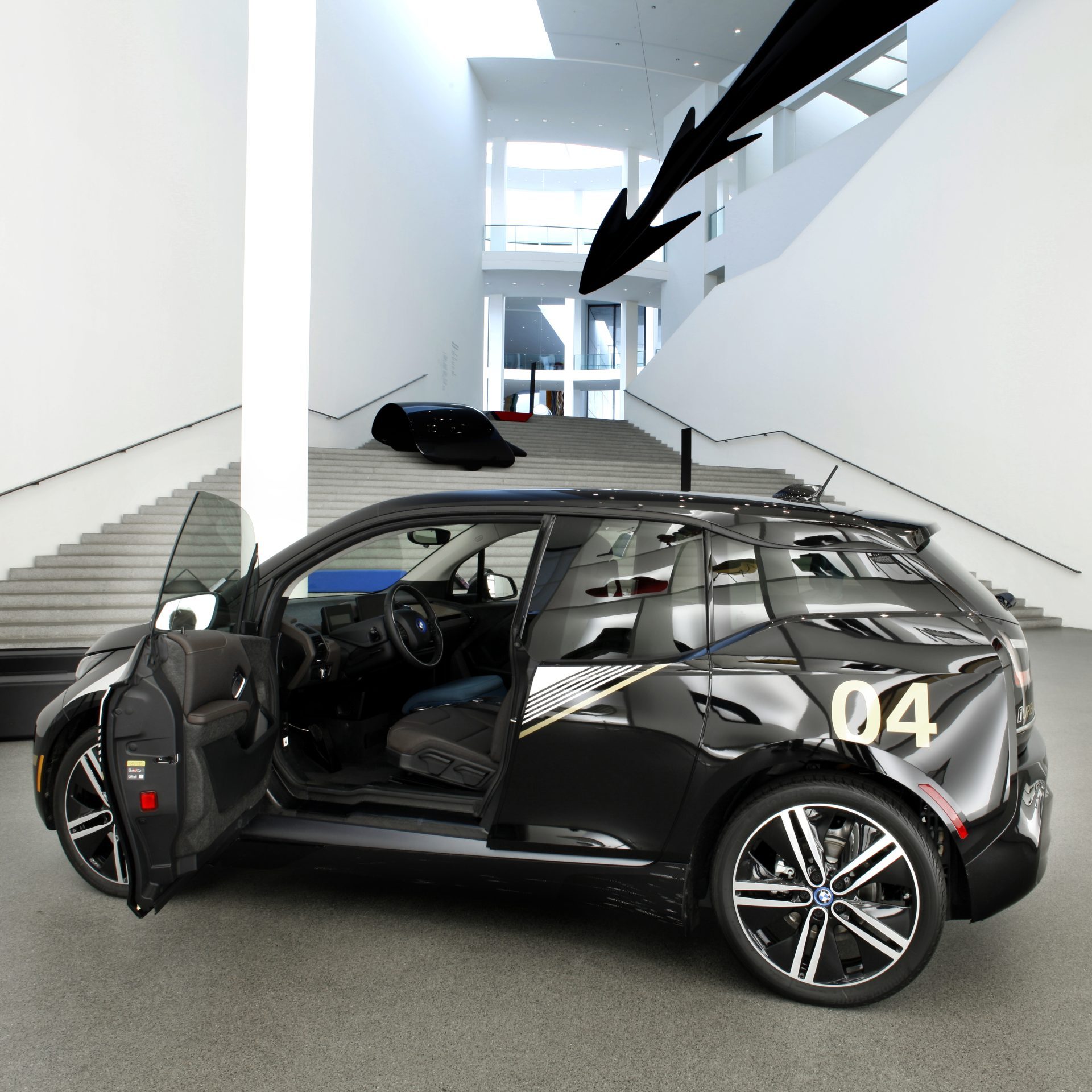 Das Foto zeigt den BMW i3 Urban Suite in Schwarz in Seitenansicht in der Pinakothek der Moderne, unterhalb der Treppen, die zur Ausstellung der Neuen Sammlung führen. Die Fahrertür ist geöffnet. Hinten links die Zahl "04".