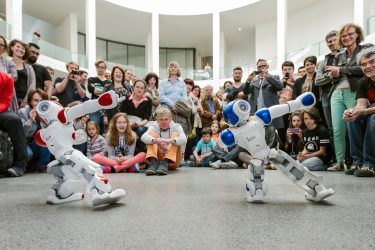 Zwei tanzende Nao-Roboter in der Rotunde der Pinakothek der Moderne. Die Roboter, weiß-rot und weiß-blau, haben jeweils einen Arm ausgestreckt. Dahinter Zuschauer:innen.