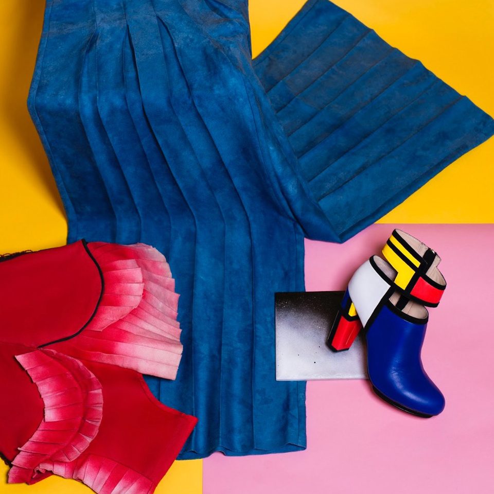 Flatlay von 2 Kleidungsstücken wie einer blauen Hose und einem Stiefel mit Absatz im Mondrian-Stil. Den Hintergrund bilden 2 orangene und 1 rosanes Viereck.