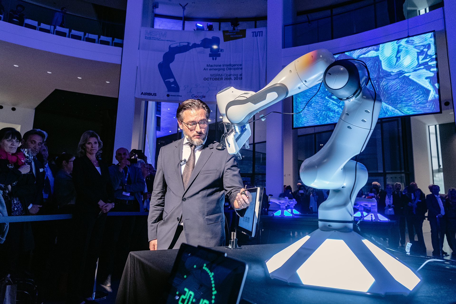 Photo of a man standing next to an industrial robot in the rotunda of the Pinakothek der Moderne. There are people and a large monitor in the background.