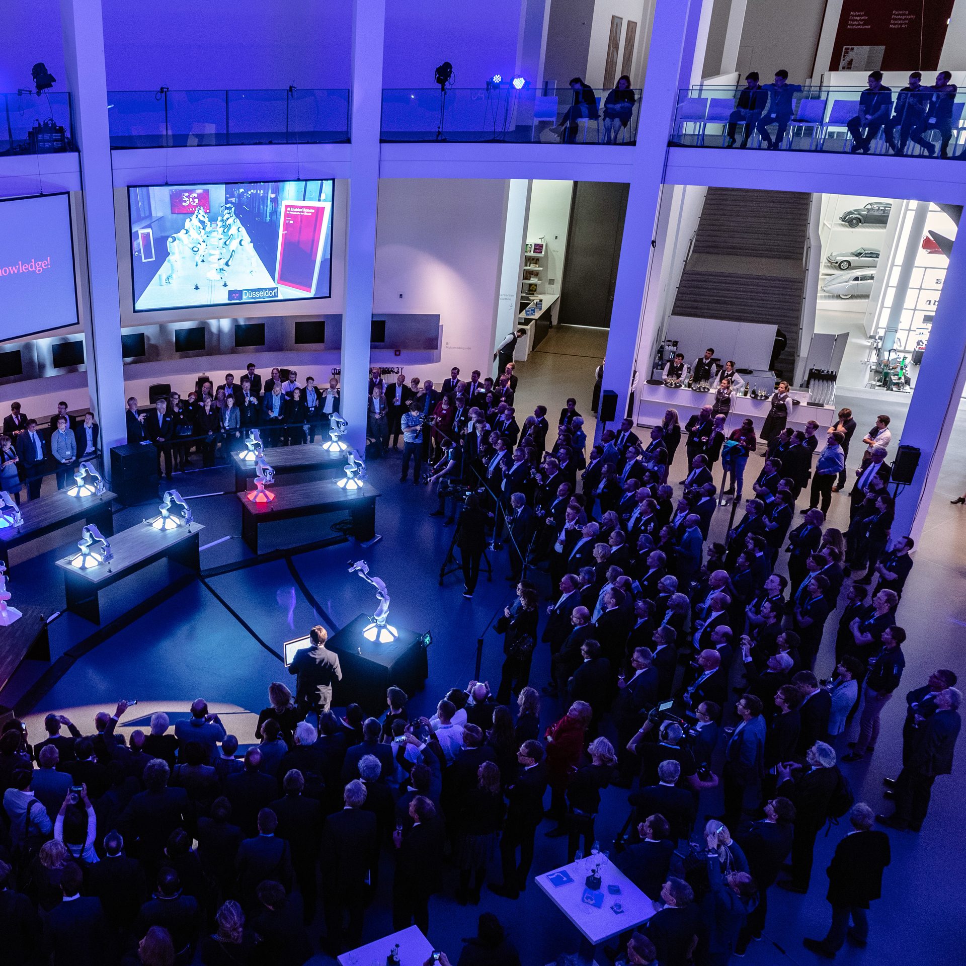 Foto von Menschen die in der Rotunde der Pinakothek der Moderne im Halbkreis um zwölf Roboterarmen stehen.