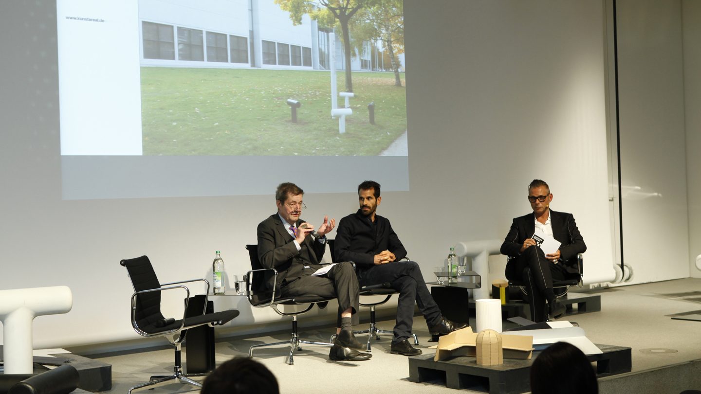 Foto von der Eröffnung. Mehrere Menschen auf der Bühne. Im Hintergrund eine Projektion mit Außenaufnahme von der Pinakothek der Moderne.