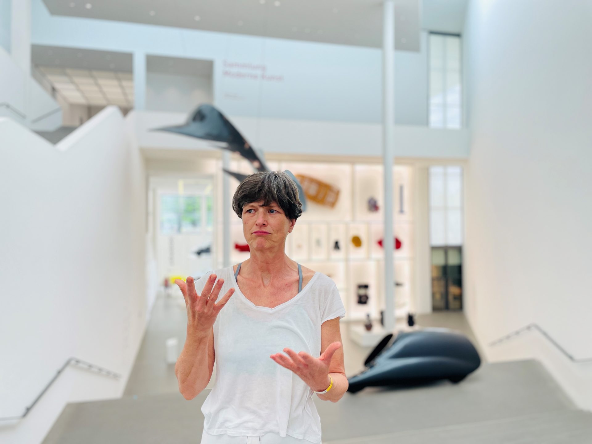 Birgit Fehn steht im Eingangsbereich des Museums und gebärdet. Sie hat kurze, graue Haare und ein weißes T-Shirt an. Im Hintergrund befindet sich der Setzkasten und zwei schwarze Objekte der Sammlung auf einer Treppe, die in das Design Museum führt.