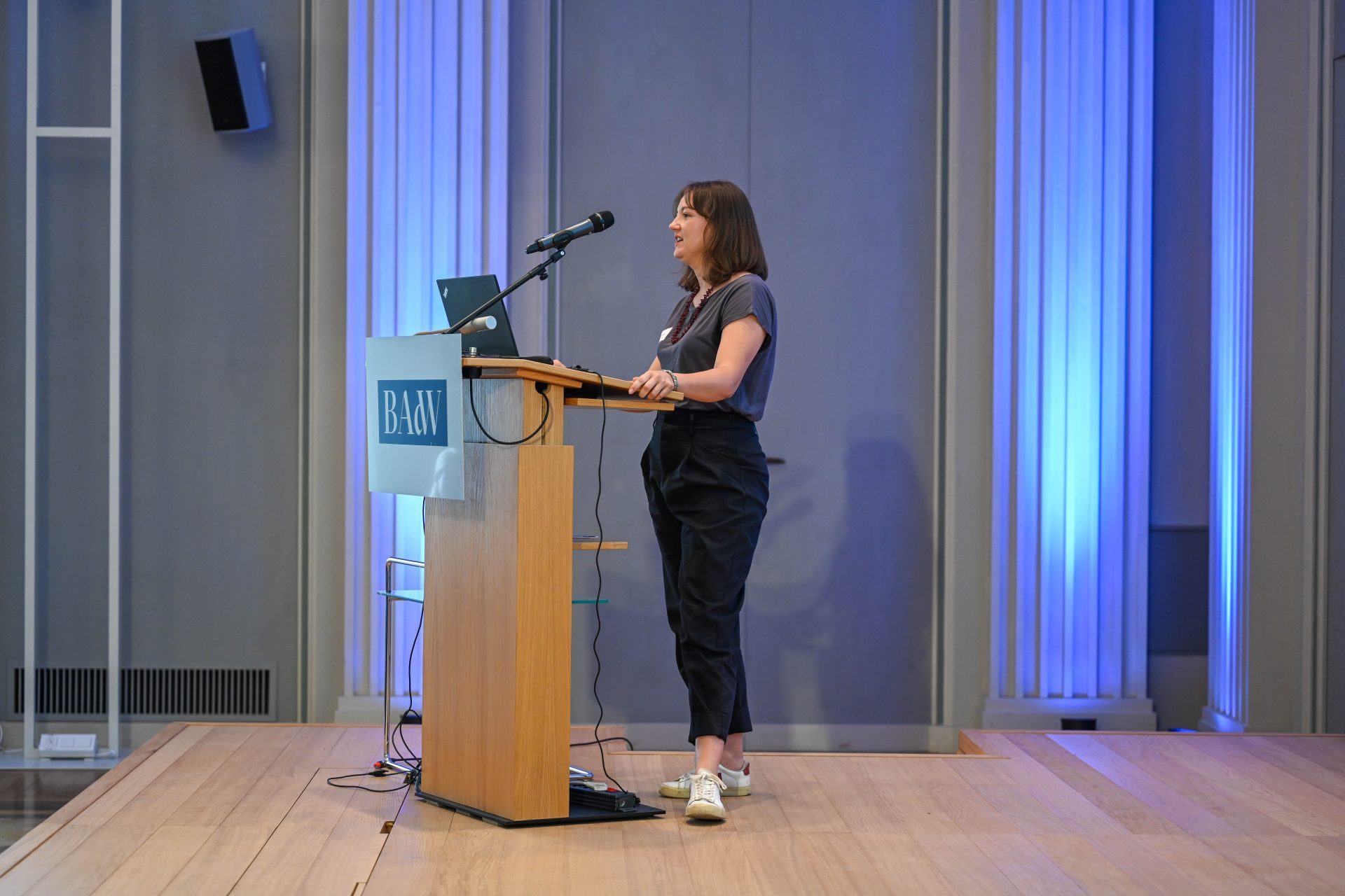 A woman with shoulder-length brown hair stands at a podium with a microphone. She is wearing black trousers and a grey T-shirt. In the background is a grey wall with blue slats in front of windows.