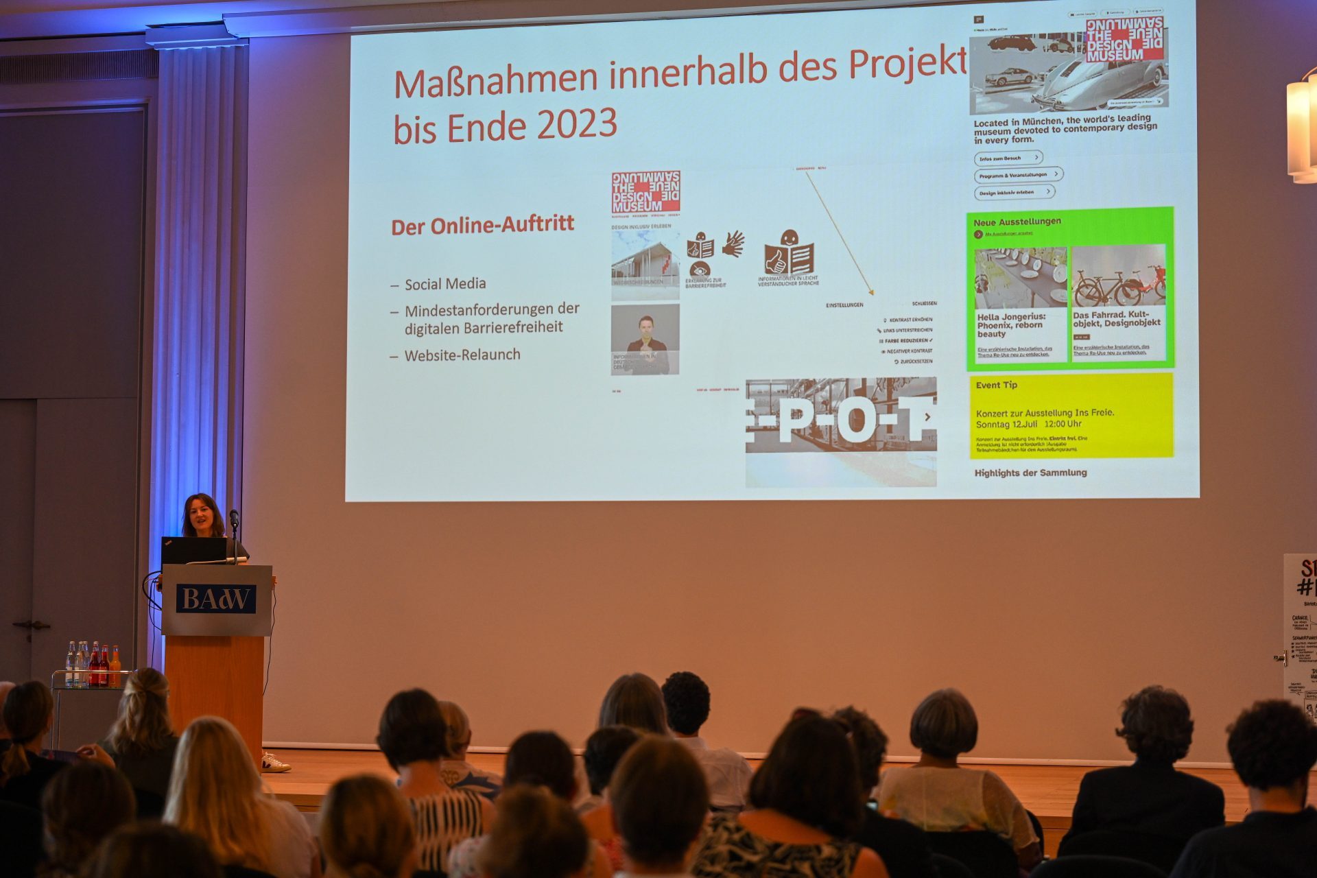 A woman stands at a podium. Behind her is a projection with the title ‘Actions within the project until the end of 2023’. Several people are sitting in the audience and their heads can be seen from behind.