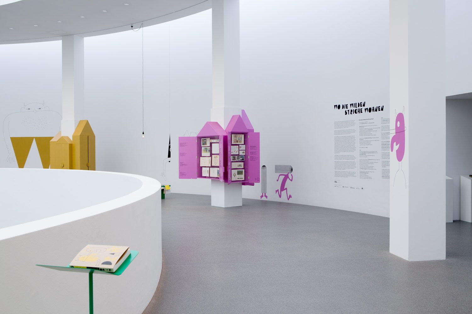 Aufnahme der Ausstellung. Man sieht farbige Schränke die Kinderbücher enthalten. An den Wänden sieht man Zeichnungen und Texte zur Ausstellung.