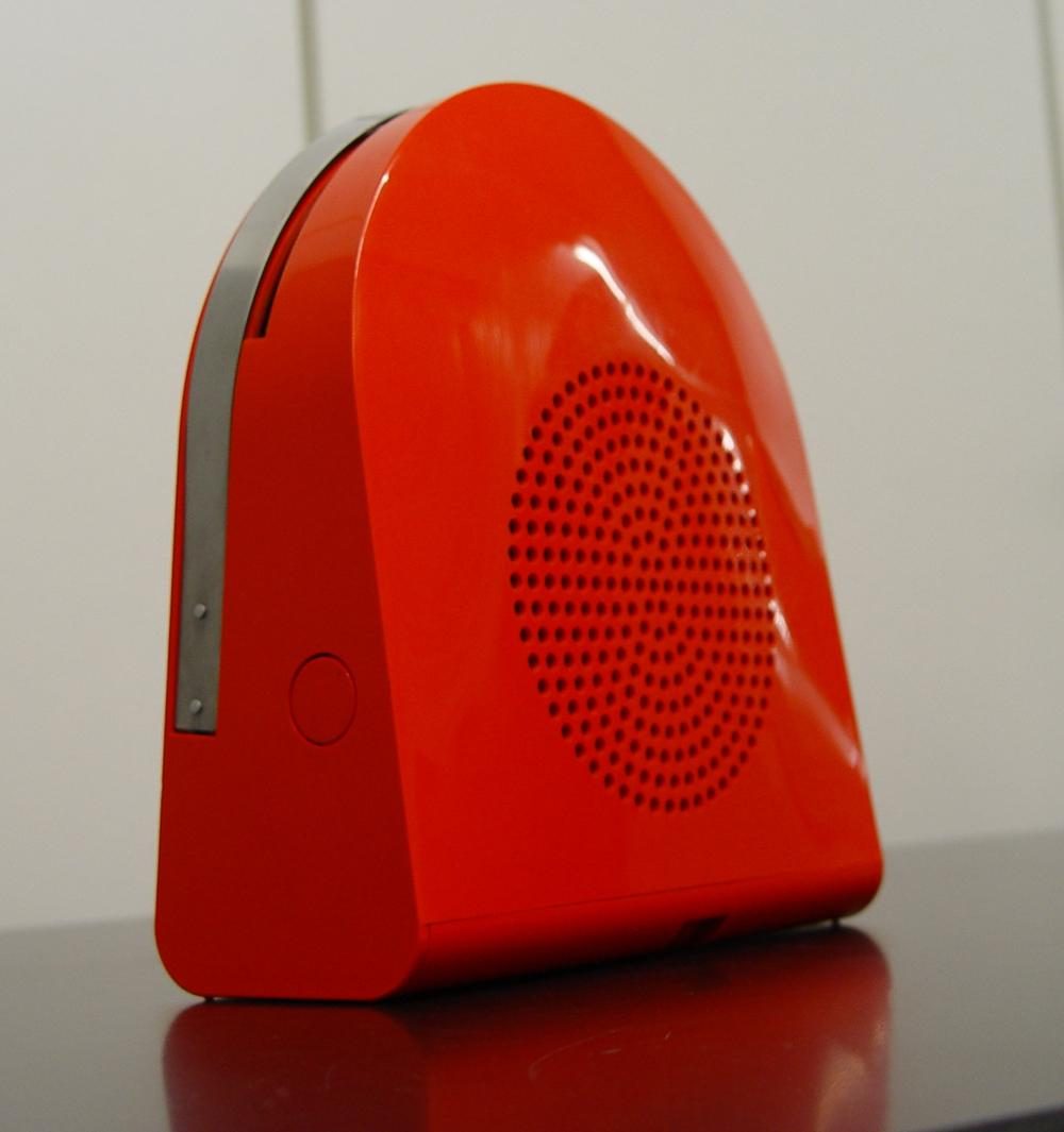 Side view of a red, portable record player with a round speaker grille and a metallic detail.