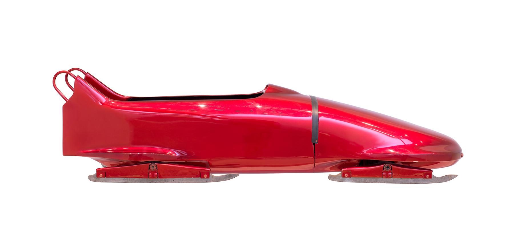 Red, aerodynamic bobsleigh with a shiny surface. The vehicle has a closed body with a narrow opening for the crew and runs on sharp metal rails.