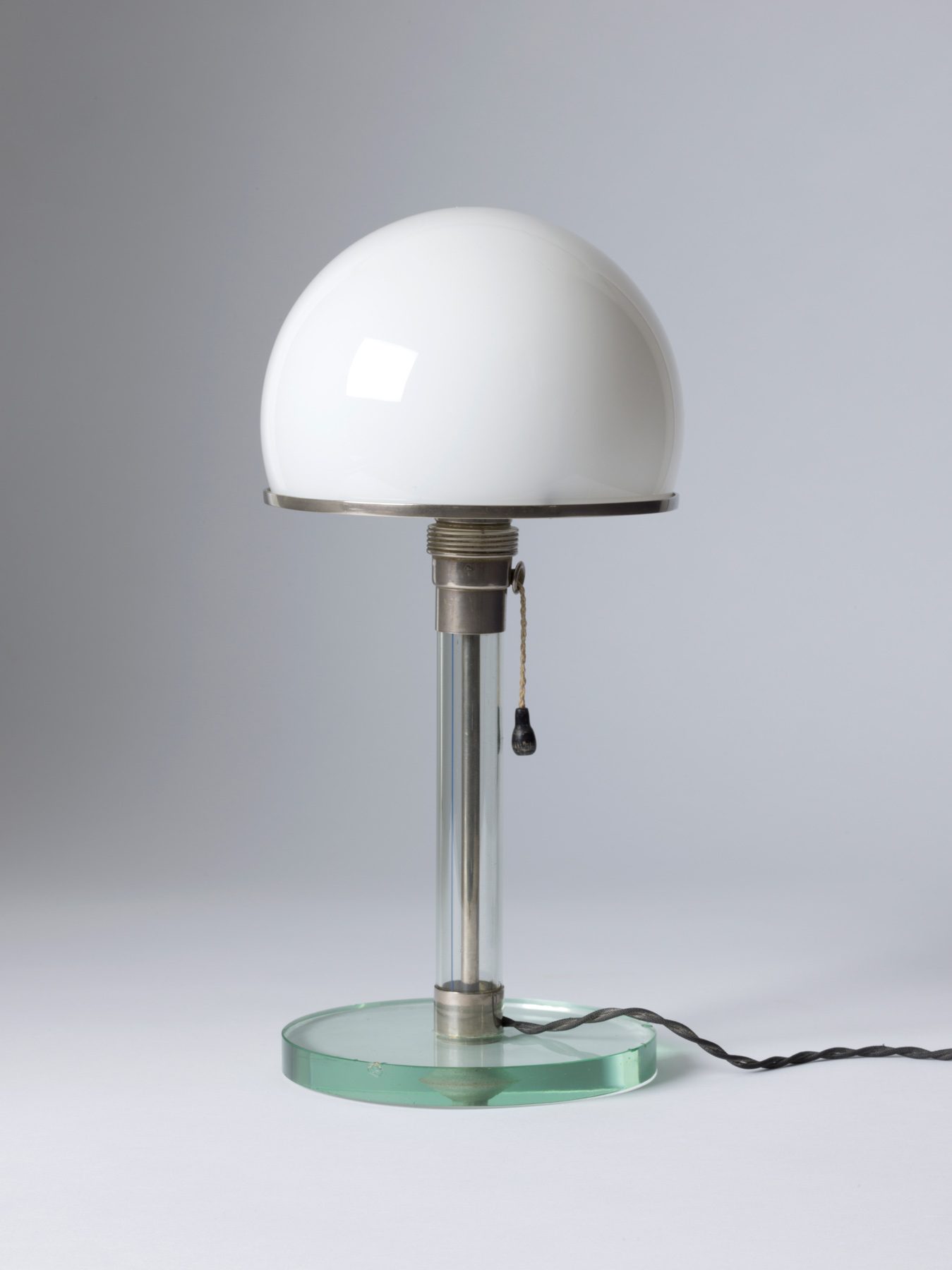 Lamp with glass base and cable in glass tube. The lampshade is a white hemisphere. The lamp has a chain as a switch that hangs down from the center. 