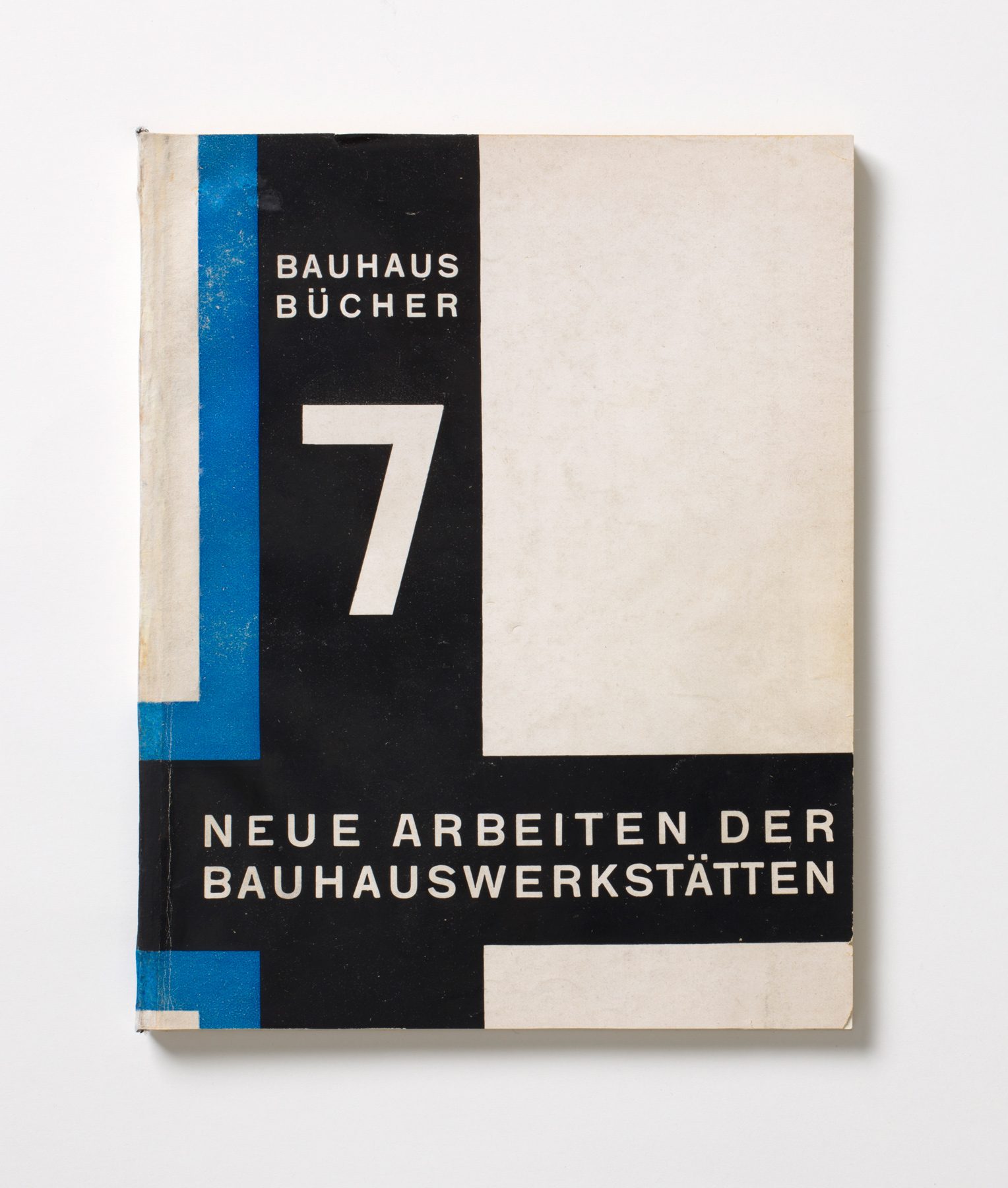 Foto Cover von Buch. Auf weißem Hintergrund bilden zwei Schwarze Streifen mit rechtem blauen Schatten ein Kreuz. Auf dem Kreuz steht in Weiß: Bauhaus Bücher, 7, Neue Arbeiten der Bauhauswerkstätten.