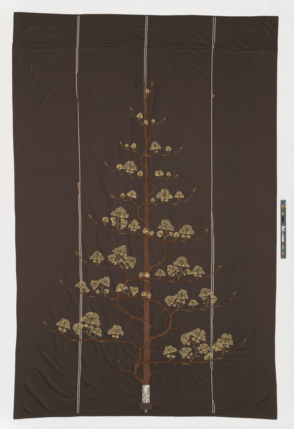 Dark brown fabric with an embroidered tree motif in the centre, whose branching branches are decorated with fine, light-coloured flowers. Three vertical lines run parallel across the fabric.