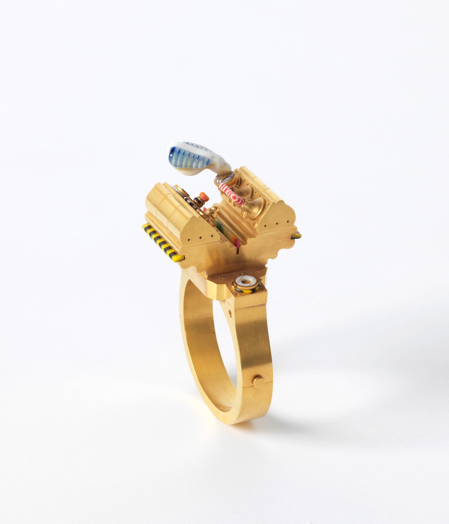 Goldfarbener Ring mit mechanischen und technischen Elementen, darunter Drähte, Rohre und eine skulpturale Verzierung, die an eine Miniaturmaschine erinnert.